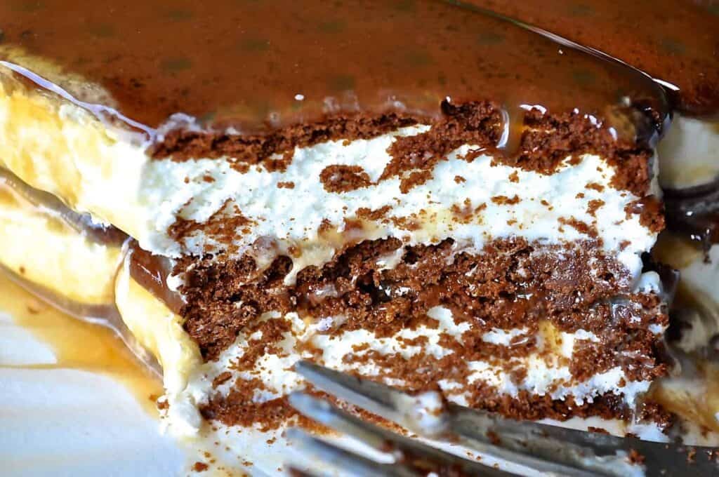 Close-up of a caramel-drizzled layered dessert with a fork, featuring cream and chocolate cake layers.