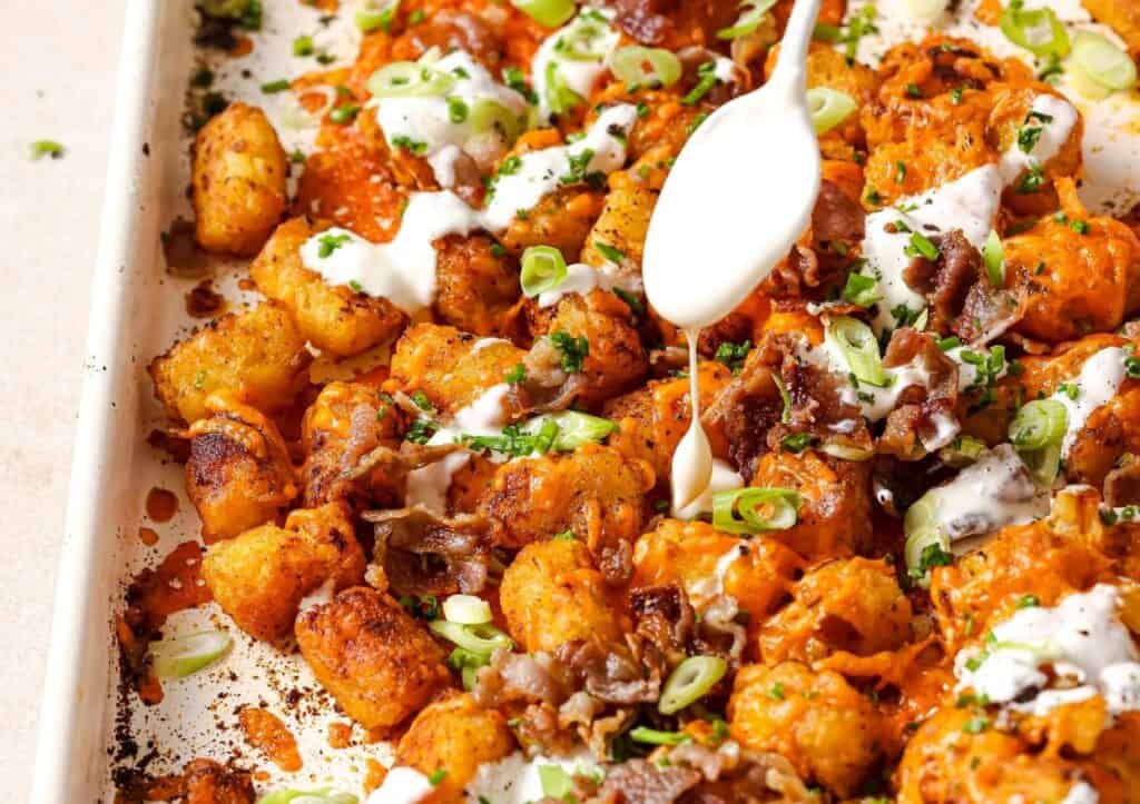 A tray of baked tater tots topped with melted cheese, green onions, chopped bacon, and drizzles of white sauce.