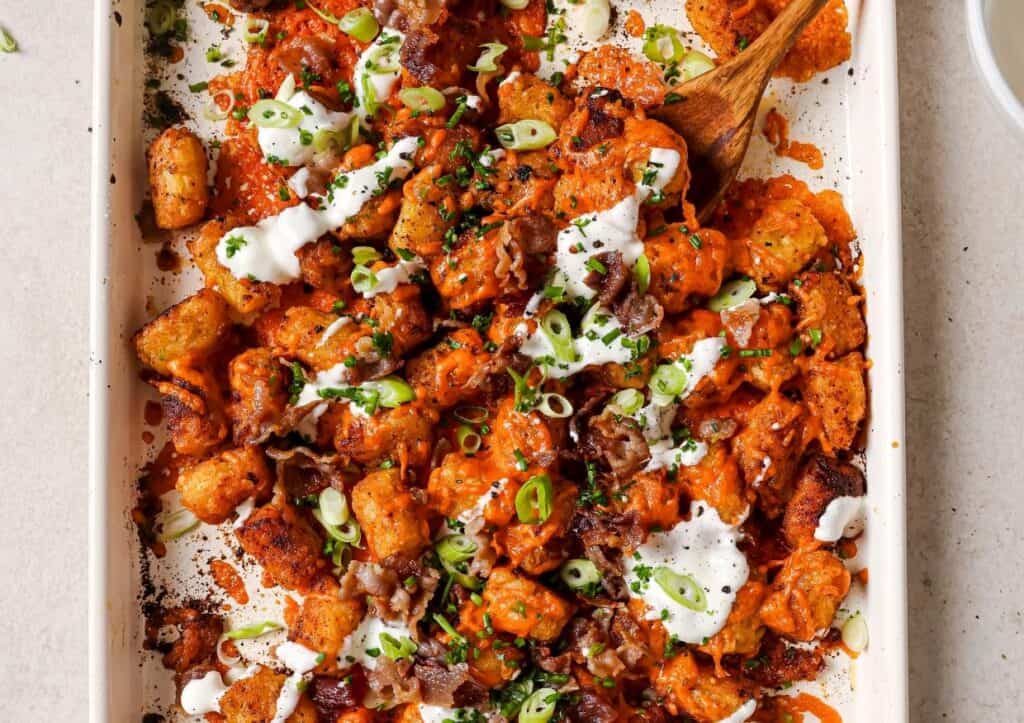 A tray of loaded tater tots topped with crumbled bacon, sliced green onions, and drizzled with a white sauce. A wooden spoon is on the tray.