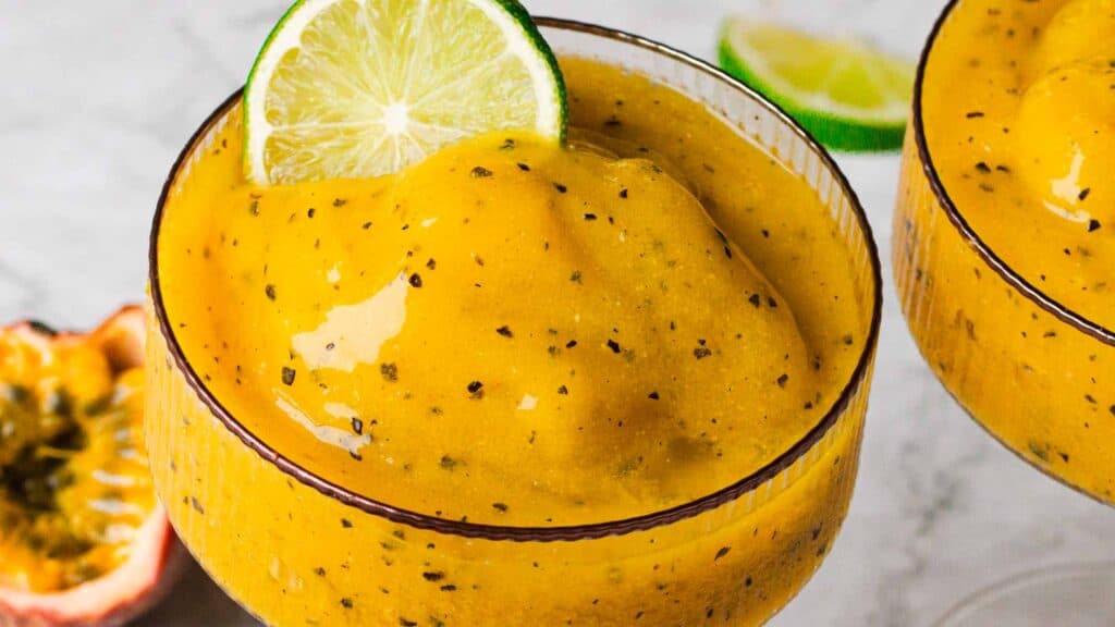 A close-up of two glasses of a yellow frozen drink garnished with lime slices, with passion fruit and a lime wedge in the background.