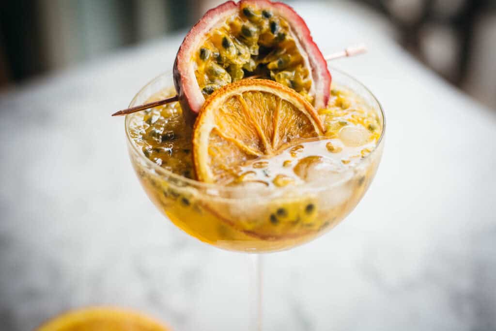 A cocktail in a glass garnished with a slice of orange and a halved passion fruit on top, containing ice and seeds. The perfect addition to your collection of summer beverages.