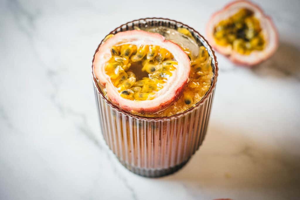 A glass with a thick rim containing a summer beverage topped with half a passion fruit showing its seeds and pulp. A second half of the passion fruit lies next to the glass.