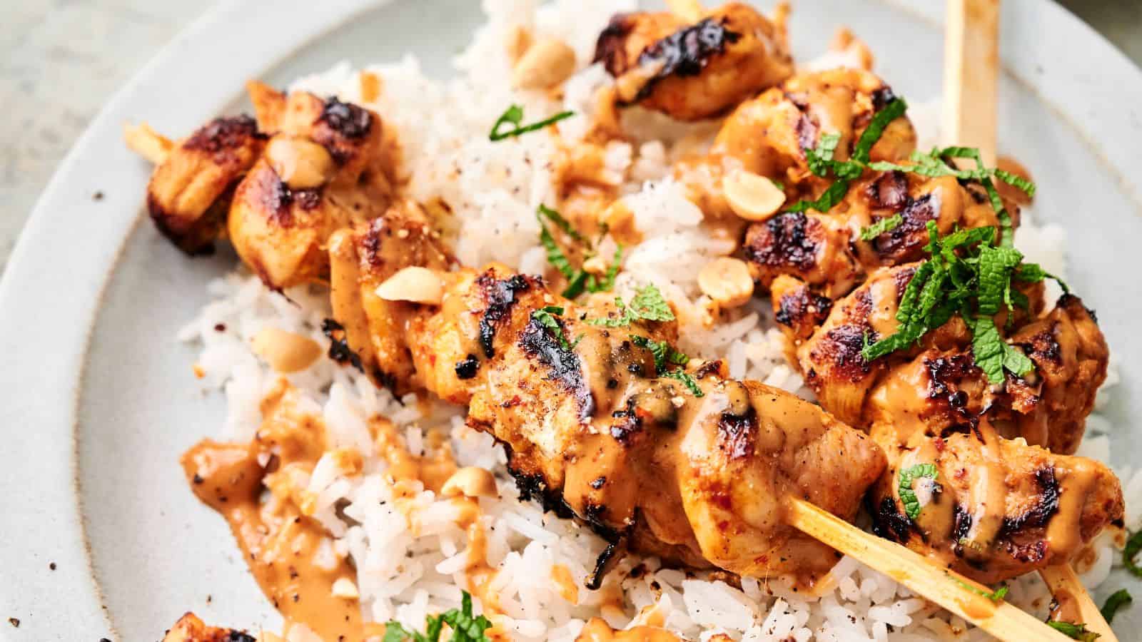 Thai chicken skewers topped with a creamy sauce and garnished with chopped herbs and peanuts, served over a bed of white rice.