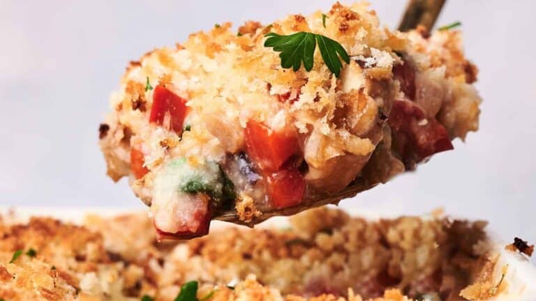 A close-up of a spoonful of vegetable casserole topped with a crispy breadcrumb crust and garnished with a parsley leaf. The casserole has visible chunks of red and green vegetables.