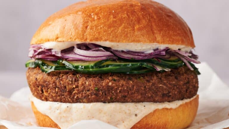 Close-up of a plant-based burger with red onions, cucumbers, and creamy sauce in a toasted bun.