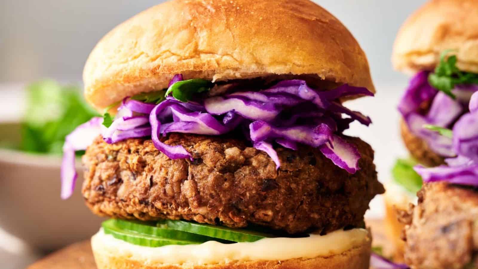 Close-up of a Veggie Burger Slider with a fried patty, sliced cucumber, shredded purple cabbage, greens, and mayonnaise in a bun. In the background, a blurred portion of another slider and a dish are visible.