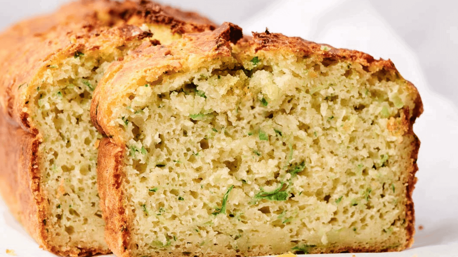 Sliced zucchini bread on a white surface, showing a moist interior with visible green flecks of zucchini.