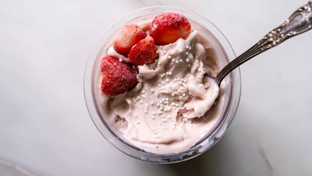 A bowl of strawberry ice cream topped with frozen strawberries and white sprinkles, with a spoon placed inside, on a white surface.