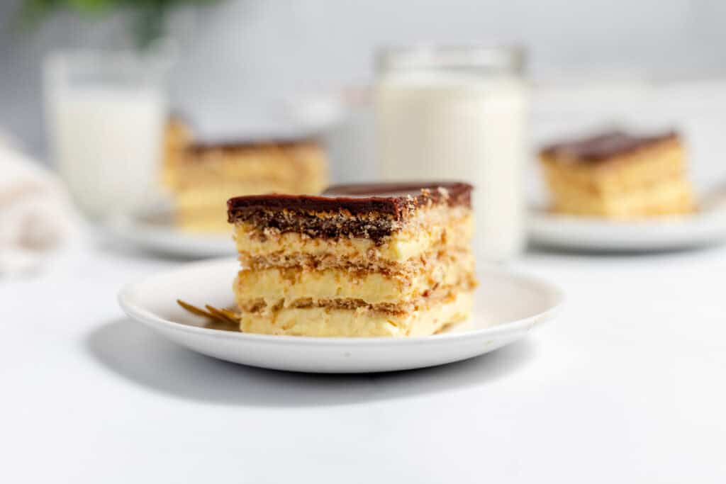 A slice of layered dessert with cream and chocolate on a white plate, with more slices and glasses of milk in the background.