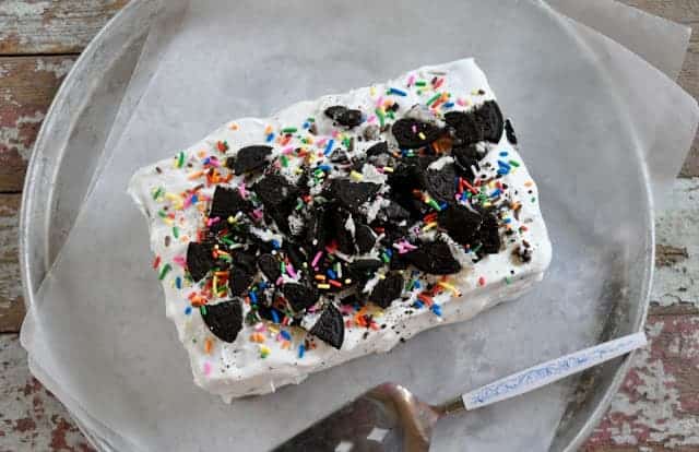 Rectangular ice cream cake topped with crumbled cookies and rainbow sprinkles, placed on parchment paper within a round metal tray. A cake server with a blue handle is visible on the right.