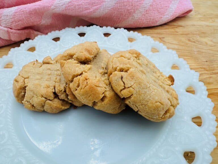 Four peanut butter cookies are neatly arranged on a white, decorative plate with a pink cloth in the background.