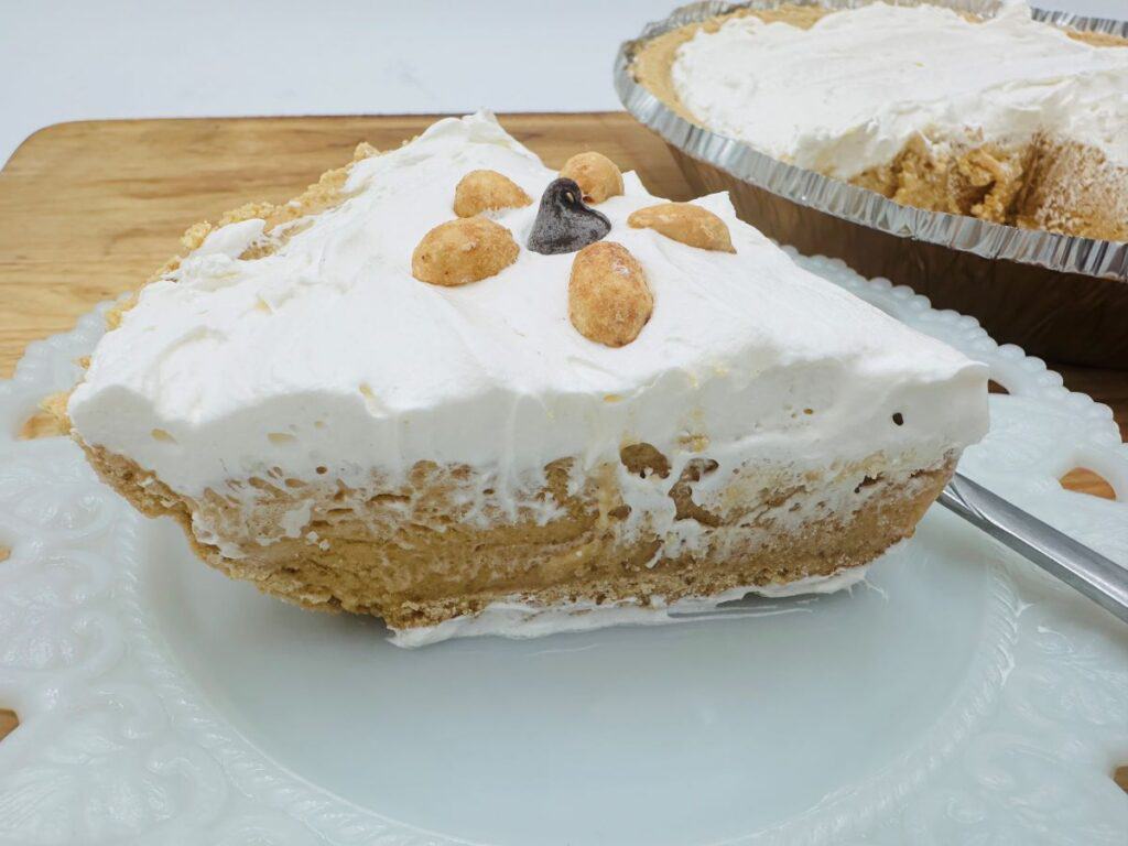 A slice of cream-topped pie with a crumbly crust, garnished with peanuts and a single chocolate chip, is served on a white textured plate. An aluminum pie dish with the remaining pie is visible in the background.