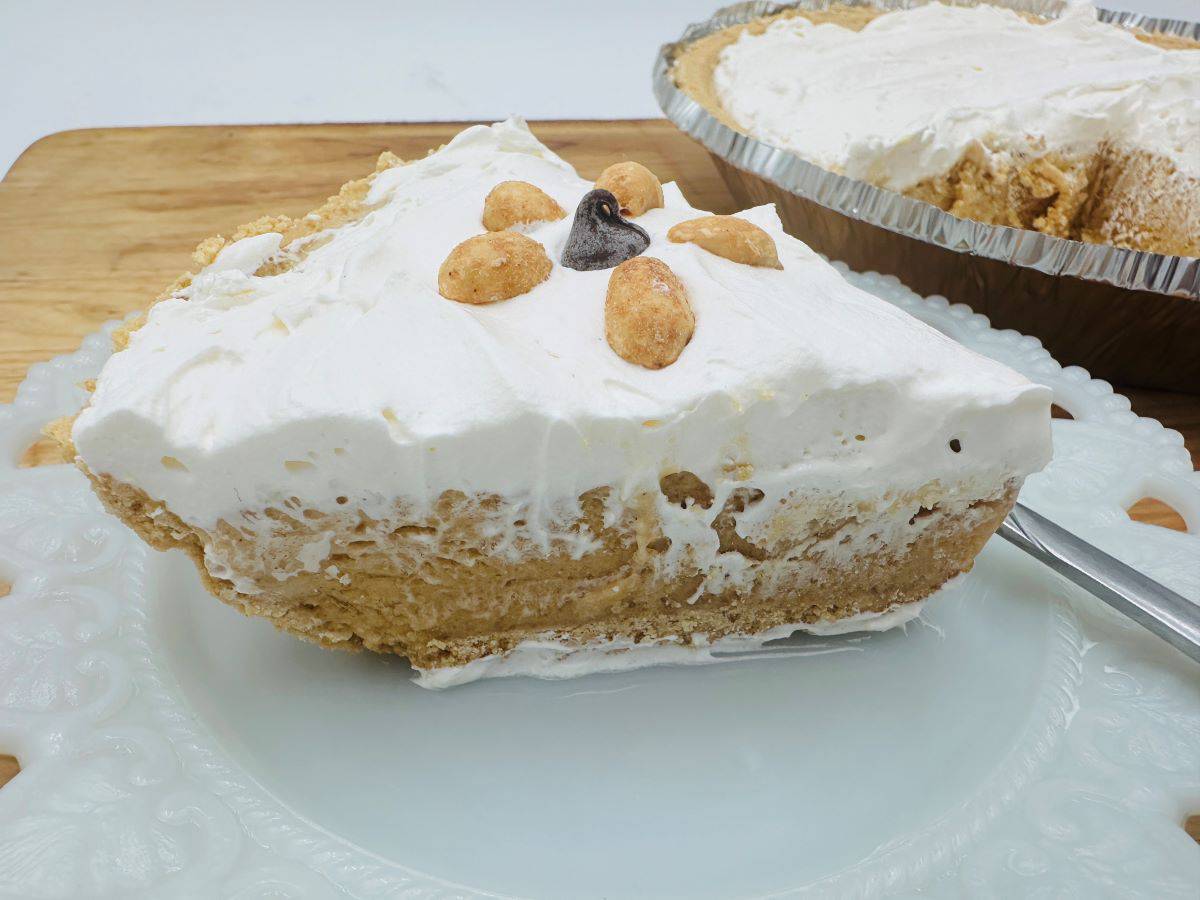 A slice of peanut butter pie topped with whipped cream and peanuts, on a white plate with the whole pie in the background.