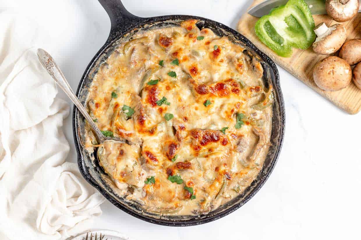 A cast iron skillet filled with baked creamy chicken and mushrooms, reminiscent of a Philly Cheesesteak Casserole, garnished with herbs, placed next to a white cloth, a fork, and a wooden board with bell pepper and mushrooms.