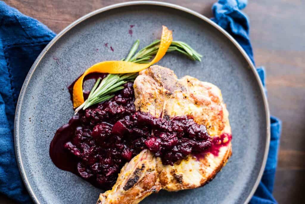 A grilled piece of chicken garnished with rosemary and orange peel, served with a dark berry sauce, presented on a light gray plate with a blue cloth napkin.