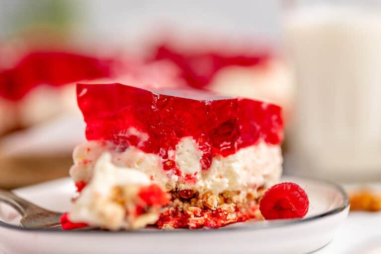 Close-up of a dessert slice featuring a red gelatin layer on top, a creamy middle layer, and a crumbly crust at the bottom, with a whole raspberry garnish on a plate.