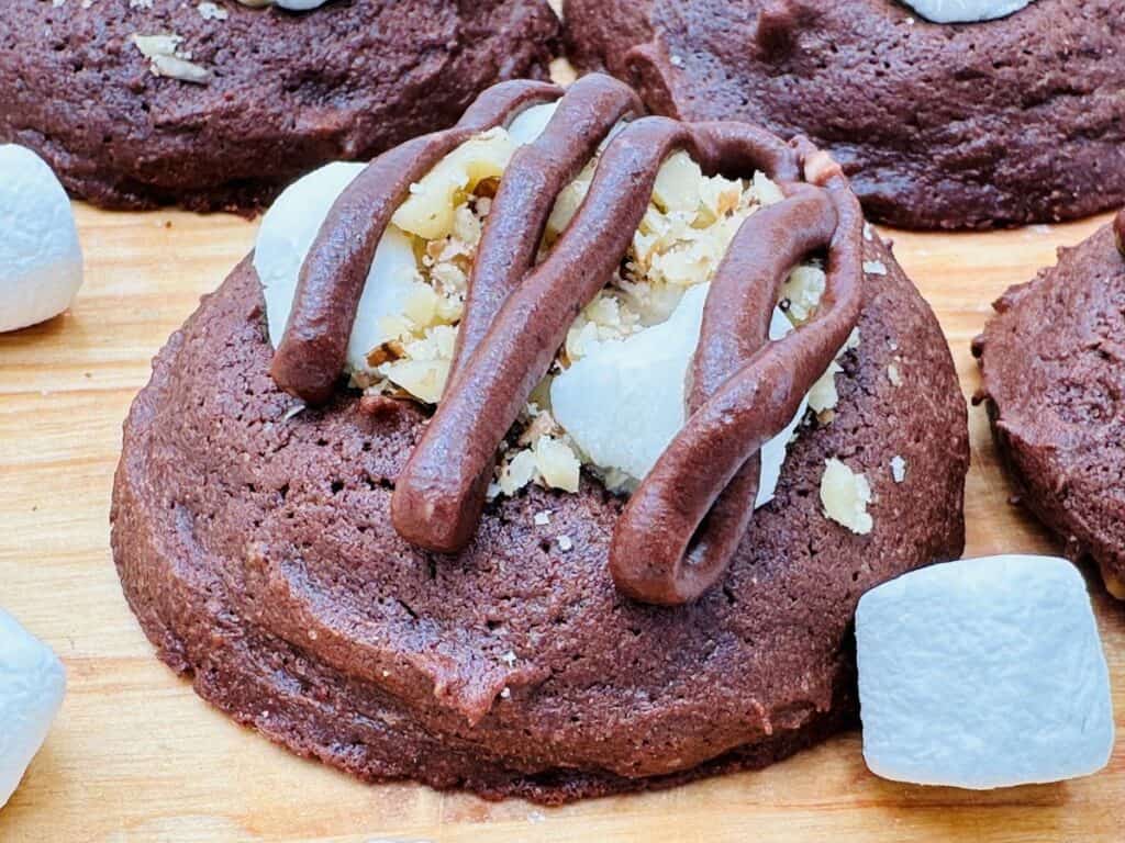 A close-up of a chocolate cookie topped with chopped nuts, marshmallow pieces, and chocolate drizzle, surrounded by additional marshmallows on a wooden surface.