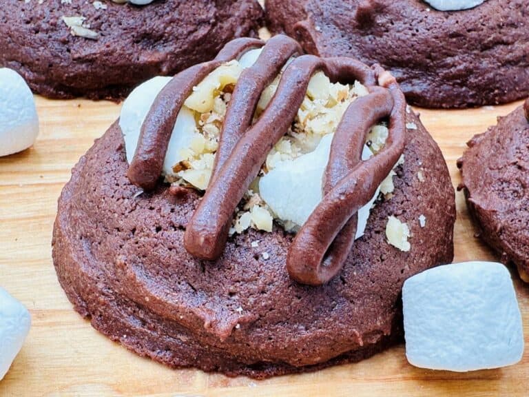 A close-up of a chocolate cookie topped with chopped nuts, marshmallow pieces, and chocolate drizzle, surrounded by additional marshmallows on a wooden surface.