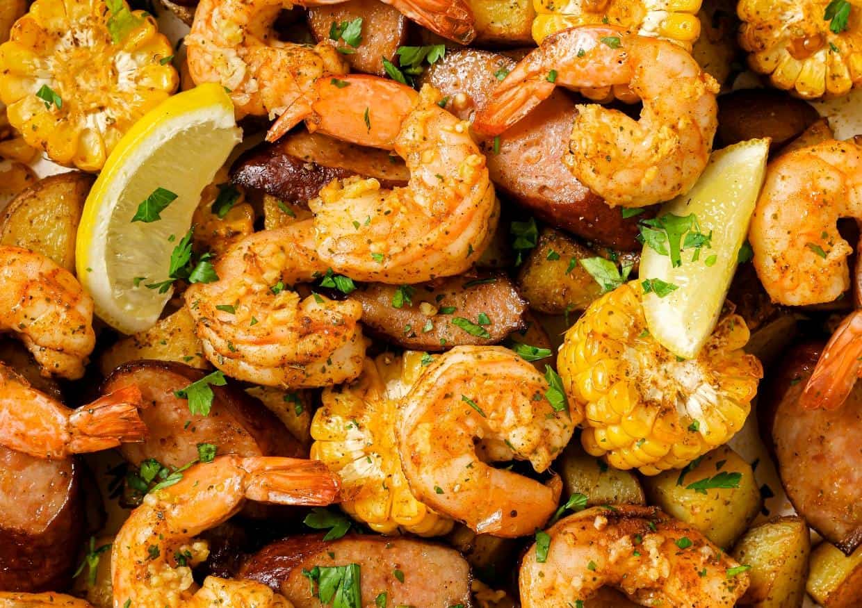 A dish of seasoned shrimp, corn on the cob, sausage slices, and potato chunks garnished with fresh herbs and lemon wedges.