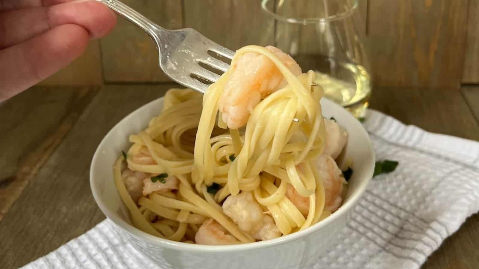 A hand holding a fork twirls spaghetti with shrimp in a white bowl, with a glass of white wine in the background on a wooden table.