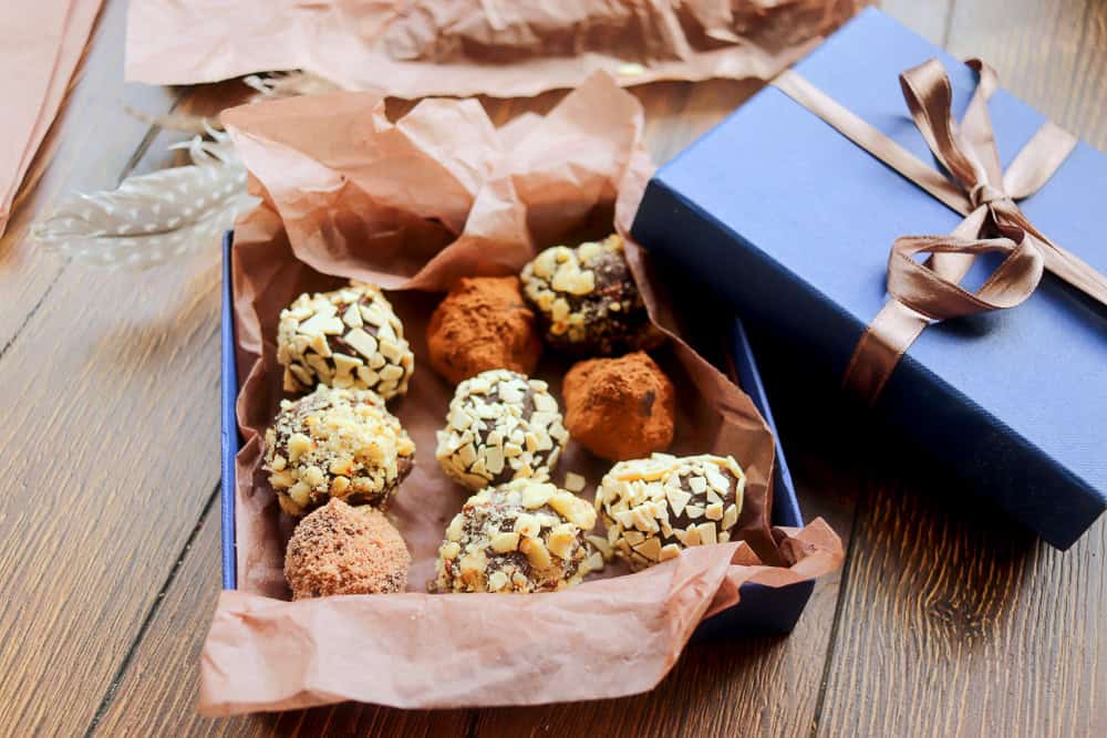 An open blue box with a ribbon contains assorted Chill Out Desserts truffles, some coated in cocoa powder and others in chopped nuts, placed on a wooden surface.