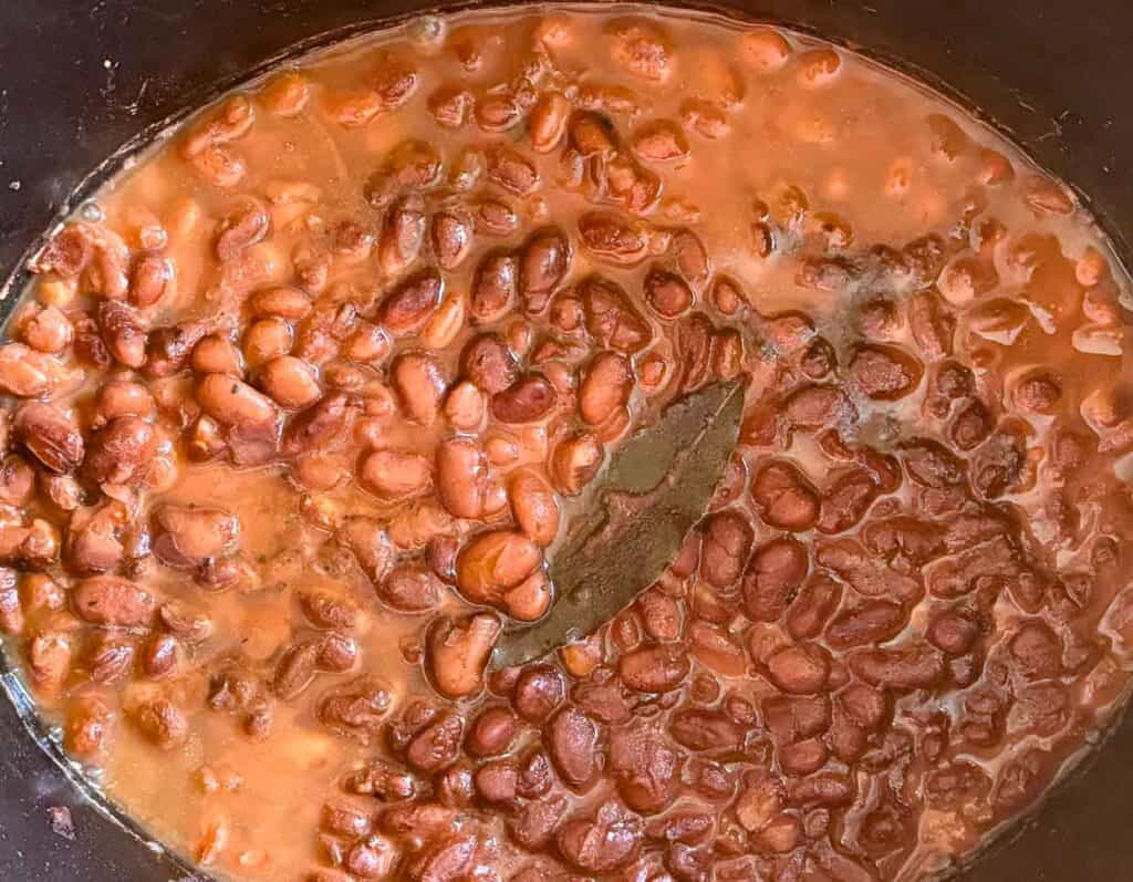 Close-up of cooked pinto beans in a thick, reddish-brown sauce in a pot. A bay leaf is visible among the beans.