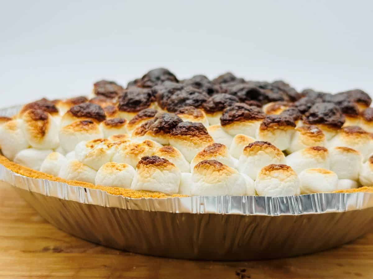 A slice of chocolate pie topped with toasted marshmallows on a white ornate plate, with a fork placed beside it, rests on a wooden surface. Another partially visible pie is in the background.