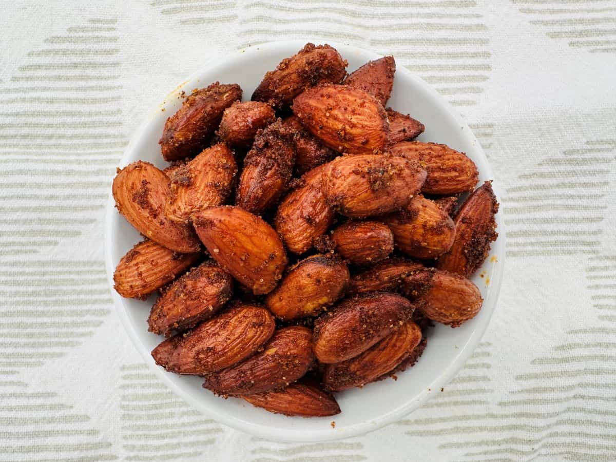 A white bowl filled with roasted, seasoned almonds is placed on a white and grey striped fabric.