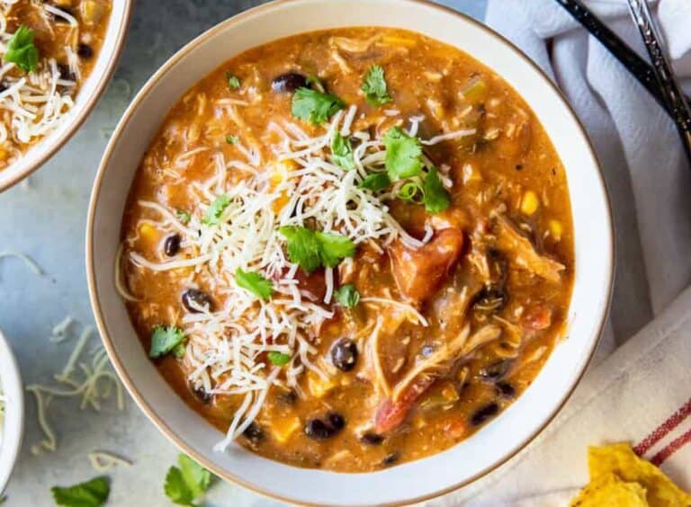 A bowl of chicken tortilla soup garnished with shredded cheese and cilantro. The soup contains black beans, corn, and tomatoes. A spoon and a napkin are visible beside the bowl.