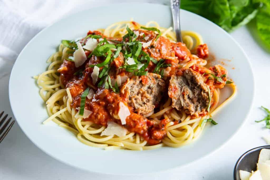 A plate of spaghetti topped with meatballs, tomato sauce, shaved Parmesan cheese, and garnished with chopped basil. A fork is placed on the right side of the dish.