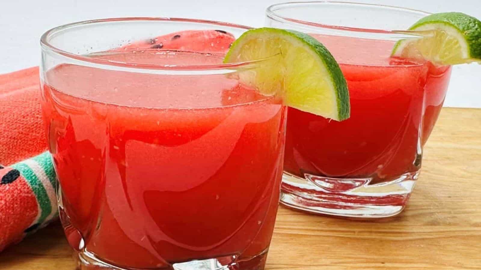 Two glasses of watermelon juice garnished with lime slices are on a wooden surface with a red napkin in the background.