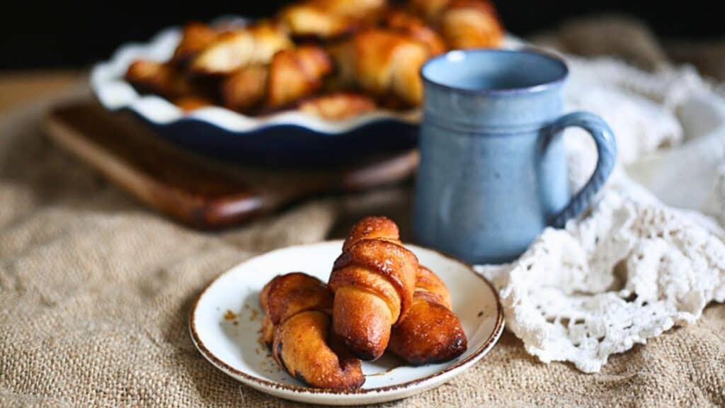 A small plate with two crescent rolls is placed in front of a blue mug and a larger dish filled with more crescent rolls, on a burlap cloth and a lace napkin in the background.