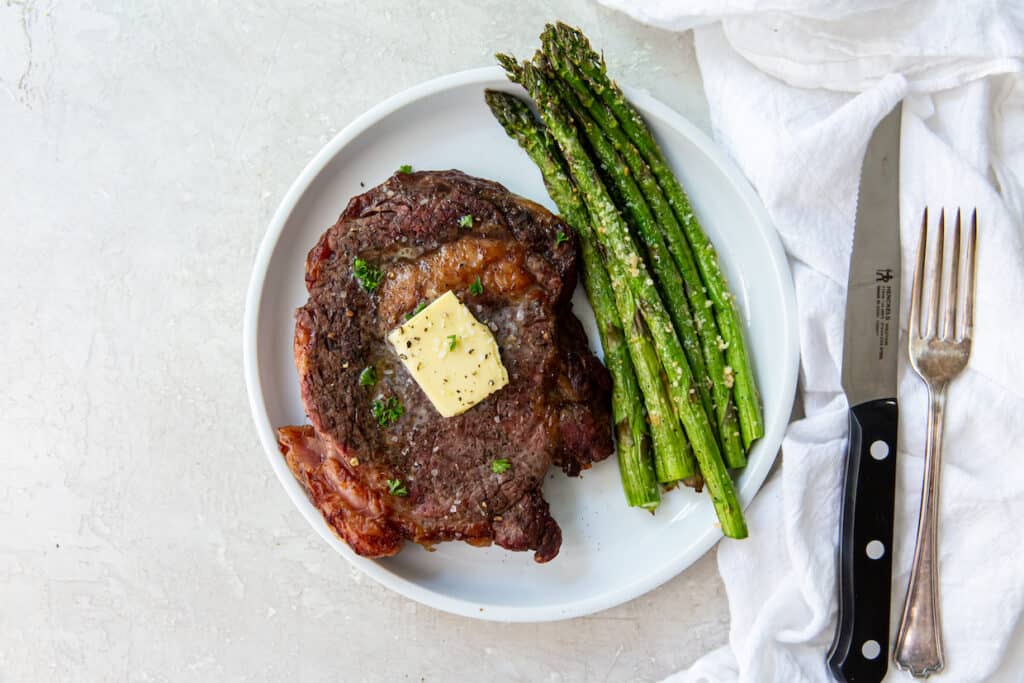 A grilled steak topped with a pat of butter is served on a white plate with a side of roasted asparagus. A knife and fork rest on a white cloth napkin beside the plate.