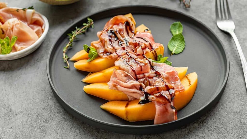 Black plate with sliced cantaloupe melon, prosciutto, fresh mint leaves, and balsamic glaze. Fork is placed on the right side of the plate. Small bowl with additional prosciutto in the background—perfect for your next BBQ party recipes collection.