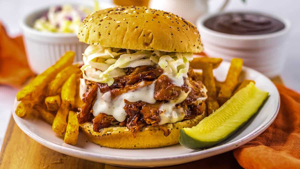 A chicken sandwich topped with coleslaw on a seeded bun, served with French fries and a pickle spear on a white plate.