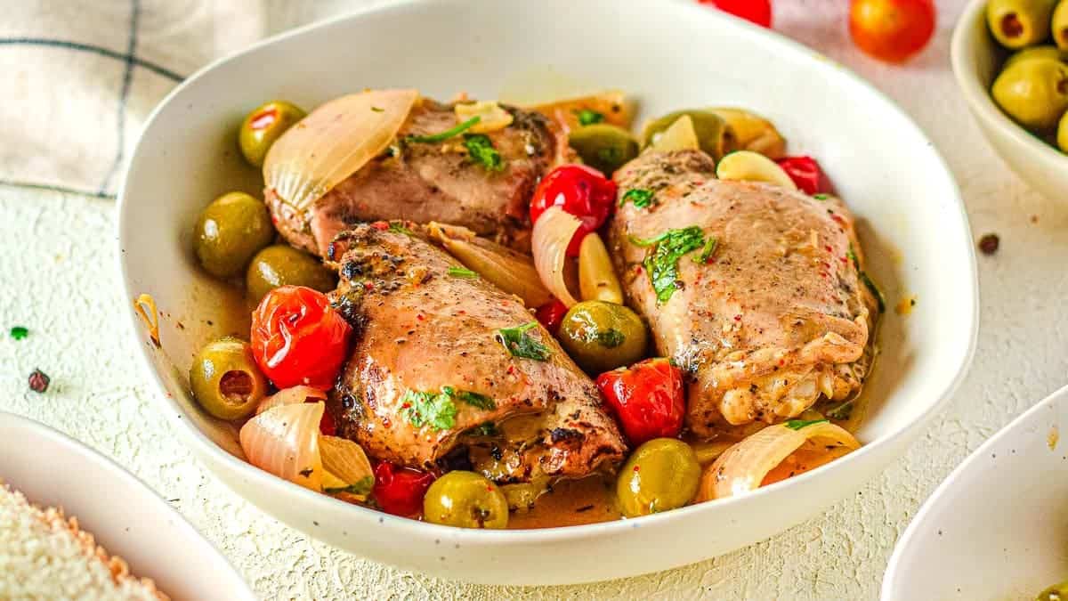 A bowl containing pieces of cooked chicken with onions, olives, cherry tomatoes, and herbs in a brothy sauce, placed on a white surface.