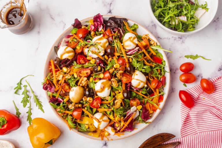 A rainbow salad featuring cherry tomatoes, carrots, greens, olives, and mozzarella balls, drizzled with dressing, served on a plate.