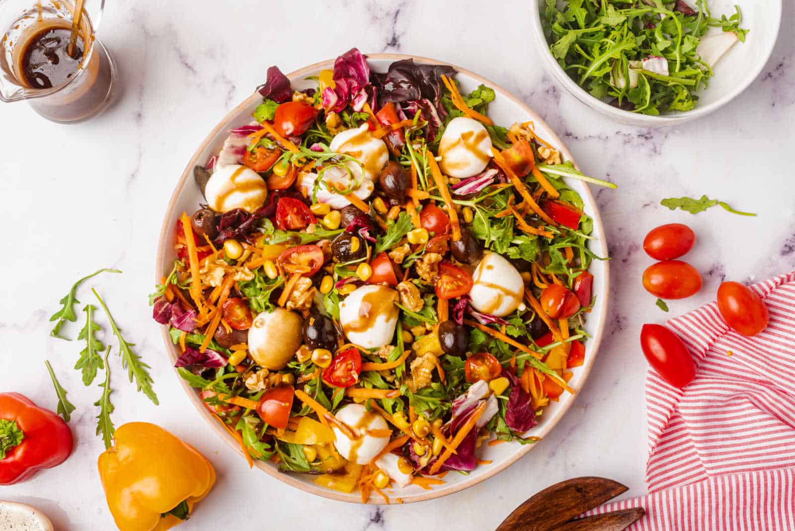 A rainbow salad with lettuce, tomatoes, carrots, olives, corn, and mozzarella balls is drizzled with dressing.