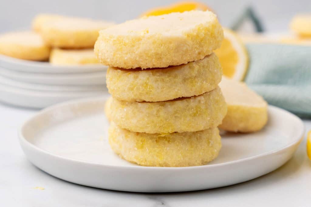 A close-up of four lemon-flavored cookies stacked on a white plate, with more cookies and a lemon in the background.