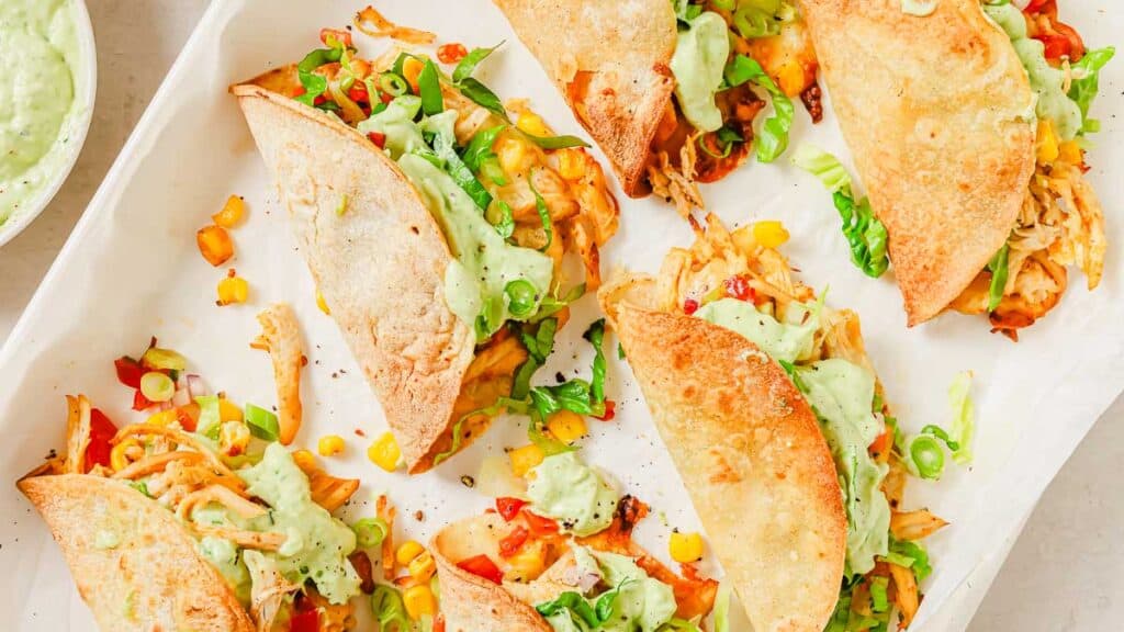 A tray of stuffed tacos filled with shredded chicken, chopped vegetables, and topped with a creamy green sauce, garnished with sliced green onions.