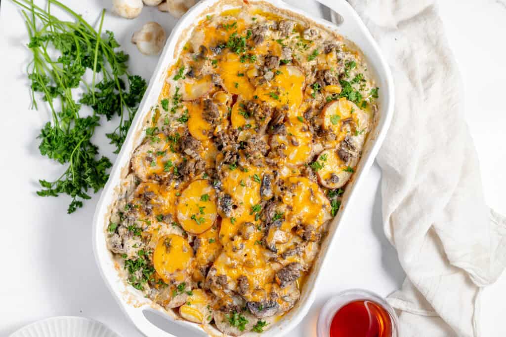 A casserole dish filled with a baked potato and cheese delight, garnished with chopped herbs, next to a bunch of fresh parsley and a small serving of red sauce. Perfect for those who want fewer dishes to clean up after a delicious meal.