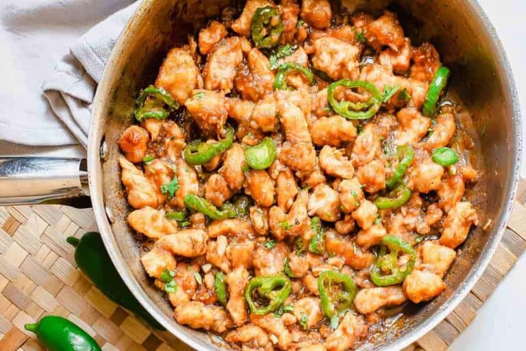 A skillet filled with cooked chicken pieces and sliced green peppers in a savory sauce. A white napkin and two whole green peppers are next to it. The skillet is on a woven placemat.