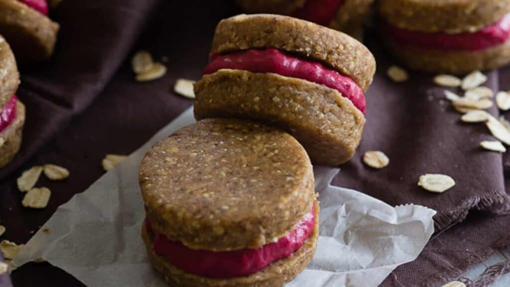 Two oatmeal cookie sandwiches filled with pink raspberry cream, placed on a piece of parchment paper, with oats scattered around.