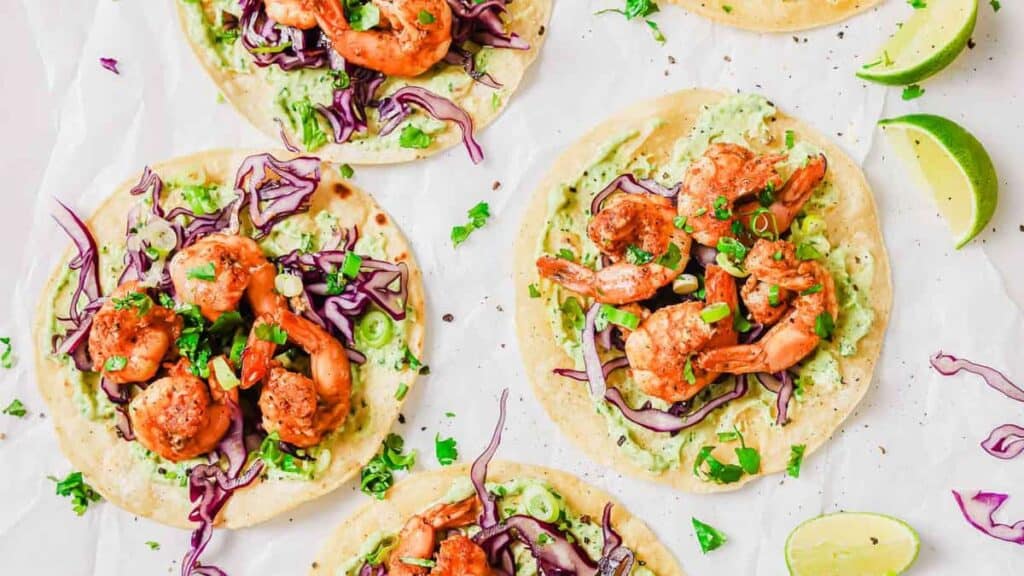 Overhead view of four shrimp tacos with avocado sauce, shredded purple cabbage, chopped green onions, and cilantro on soft tortillas. Two lime wedges are on the side.