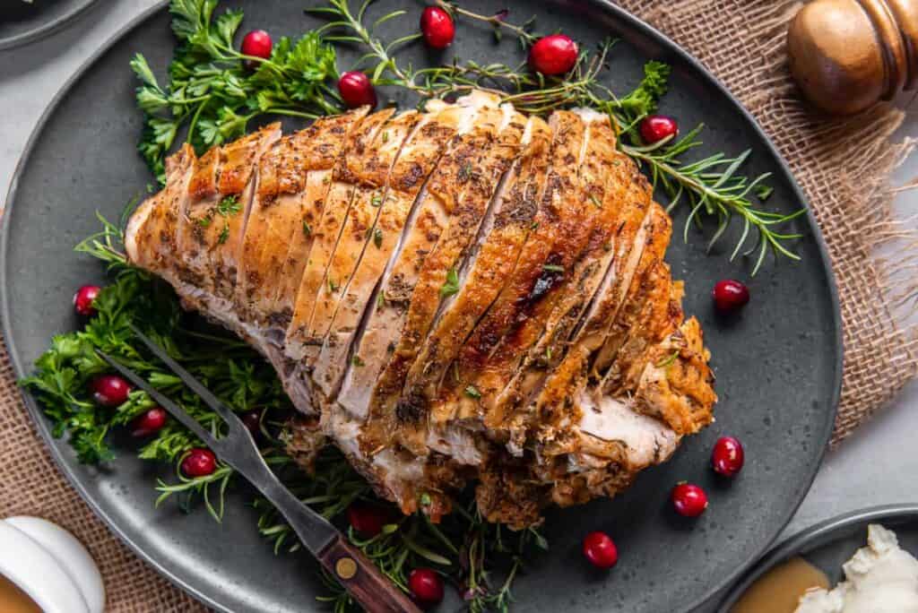 Sliced roasted turkey breast garnished with fresh herbs and cranberries on a black plate with a fork to the side.