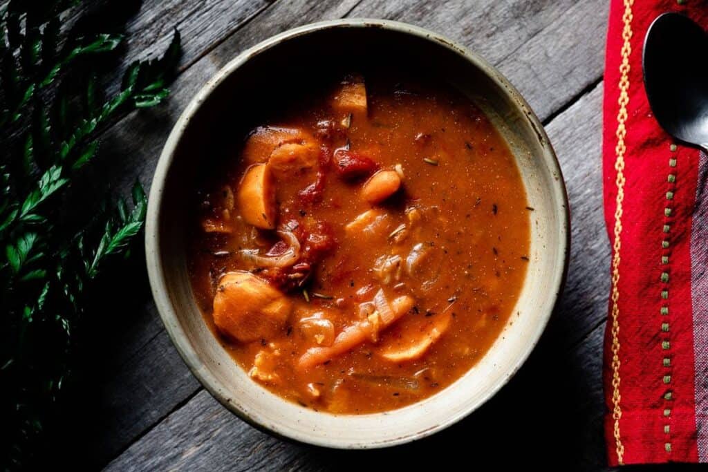 Top view of a bowl filled with a rustic tomato-based stew, with visible chunks of vegetables and herbs, placed on a wooden surface next to a red napkin and a spoon.