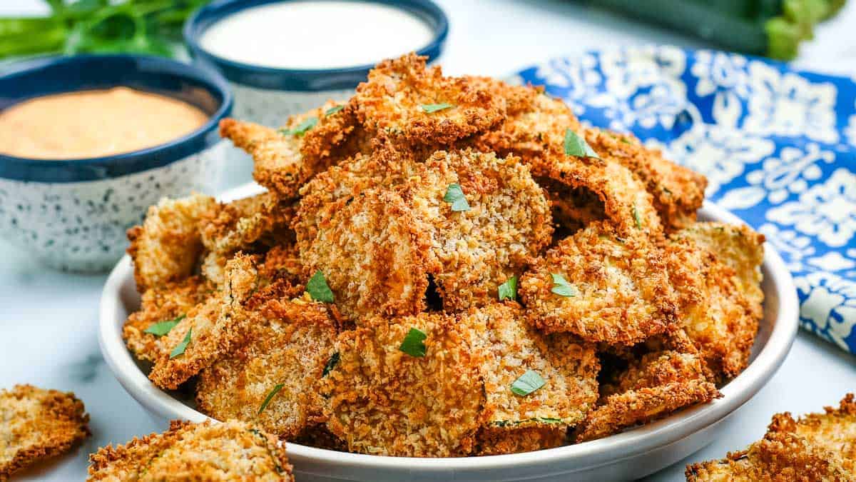 Bowl of zucchini chips with parsley in front of two bowls of dipping sauce.