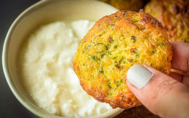 A hand holding a fried vegetable patty in front of a bowl of white dipping sauce.