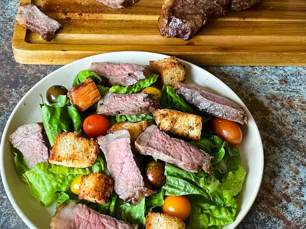 A bowl of steak salad with cherry tomatoes, romaine lettuce, and croutons. Sliced steak is placed on top of the salad, with extra steak pieces on a wooden cutting board in the background.