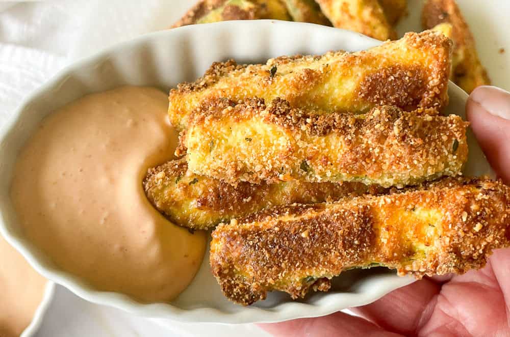 A small white dish holds several crispy zucchini sticks next to a creamy dipping sauce. A hand is partially visible, holding the dish.